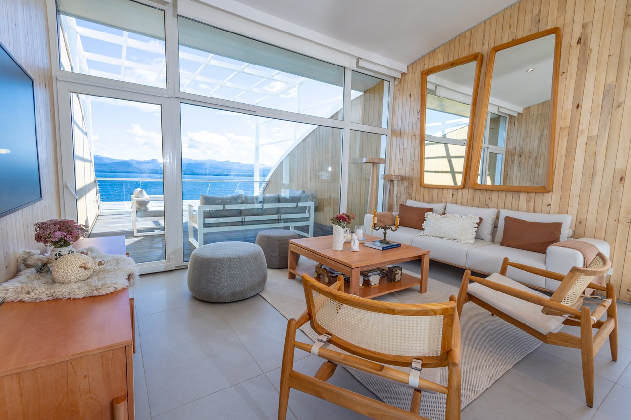 Living room with a white sofa and woven chairs, opening to a balcony with water and mountain views.