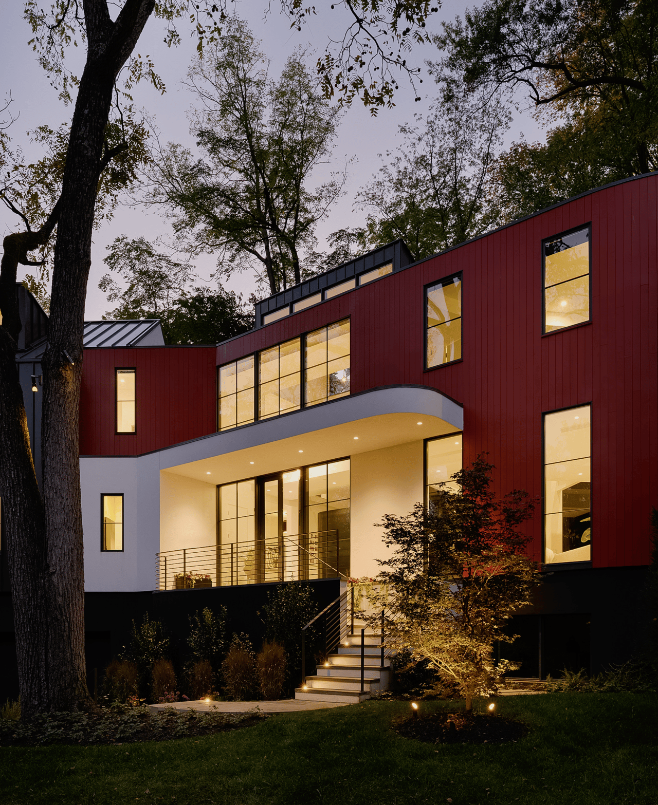 Modern red-sided home glowing at dusk, framed by mature trees and landscaped steps.