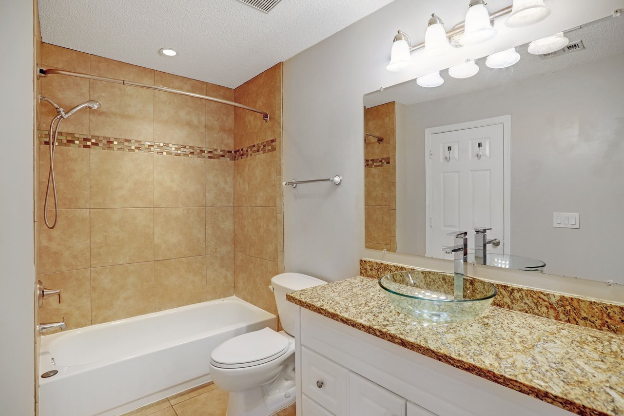 Bathroom with a tan tiled tub surround and granite vanity with a glass vessel sink.