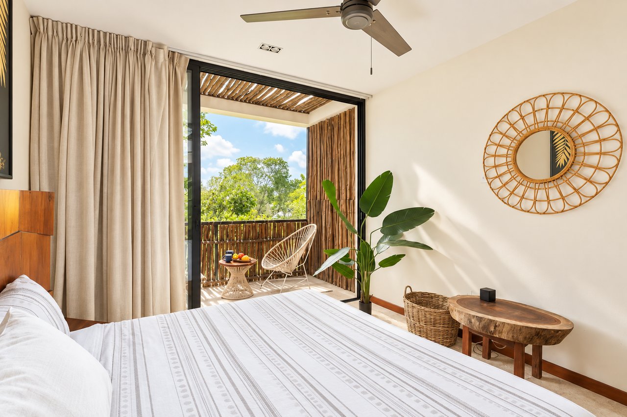 Furnished bedroom opening onto a sunny balcony with woven chairs and leafy greenery.