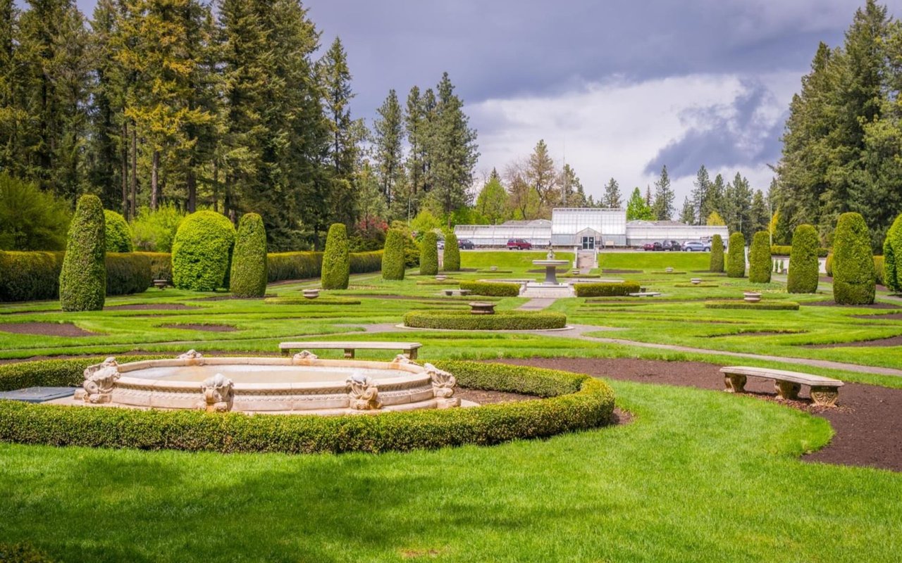 Formal garden with a stone fountain, clipped hedges, paths, and a greenhouse in the distance.