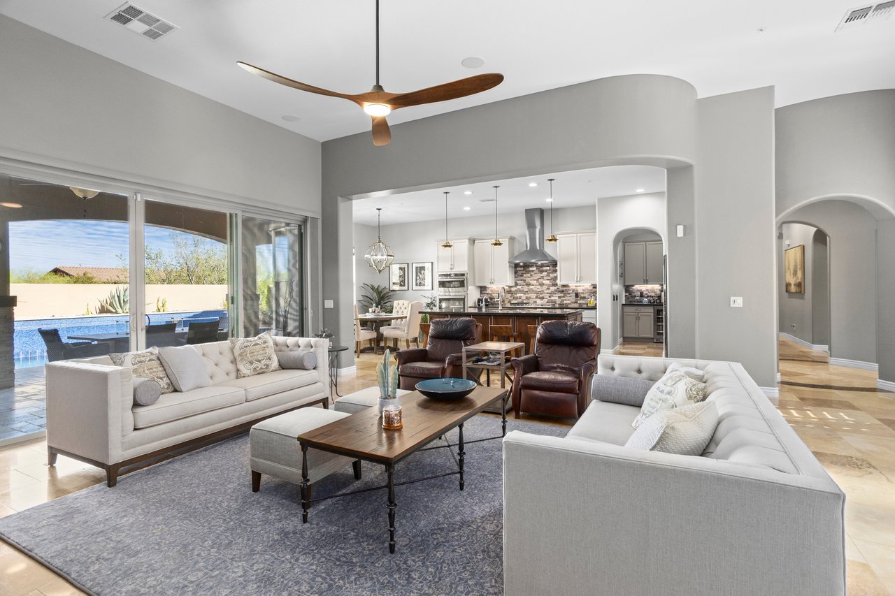Bright staged living room with two sofas, recliners, coffee tables, and a view into the kitchen and out to a patio and pool through large sliding doors.