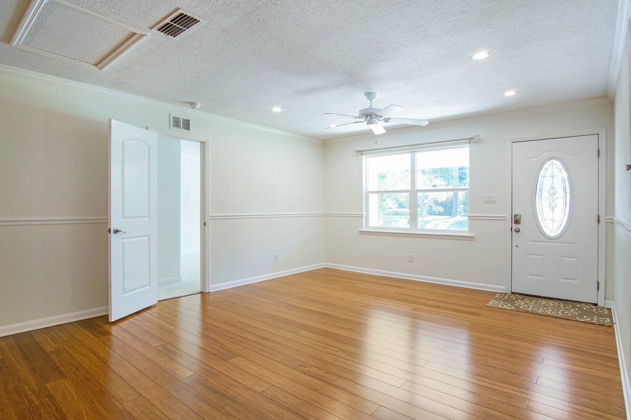 A bright, empty living room with wooden floors, beige walls, a ceiling fan, and recessed lighting. A large window and oval glass door let in sunlight.