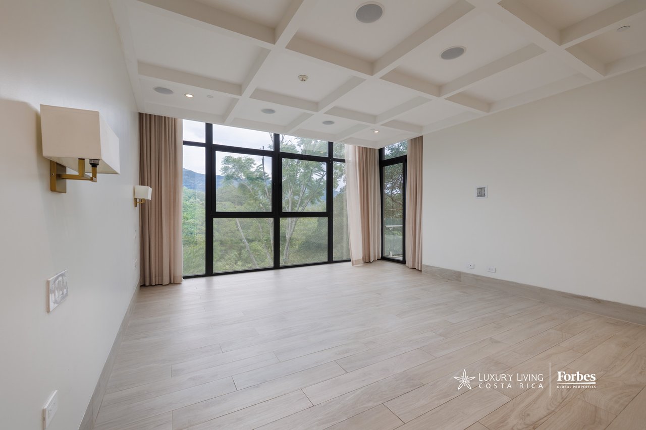 Vacant apartment room with coffered ceiling, large windows, and views of trees and distant hills.
