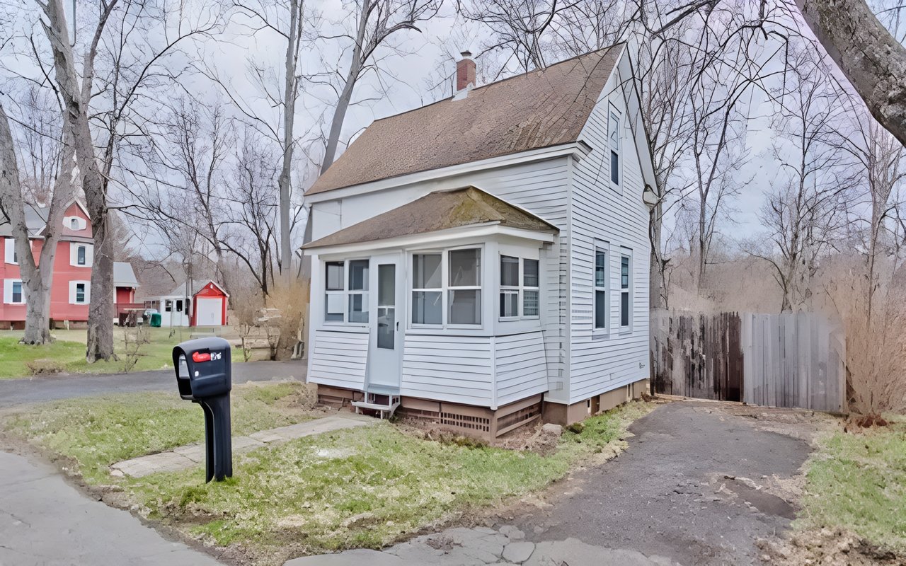 White two-story house with an enclosed porch, steep roof, and driveway beside a black mailbox.