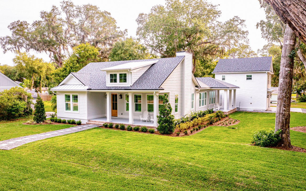 White home with a covered porch and gray roof on a broad lawn beneath mature moss-draped trees.
