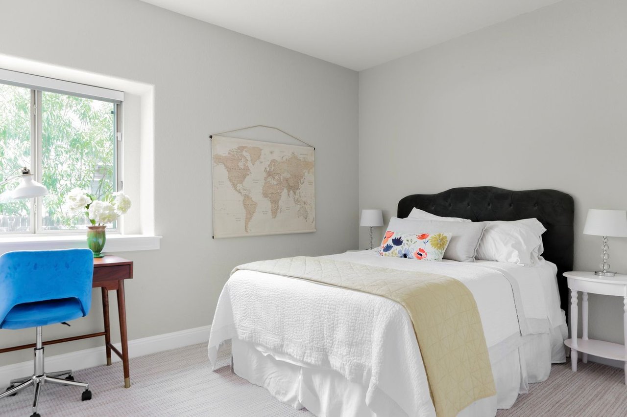 Bright bedroom with a white bed, black tufted headboard, and blue desk chair beside a window.