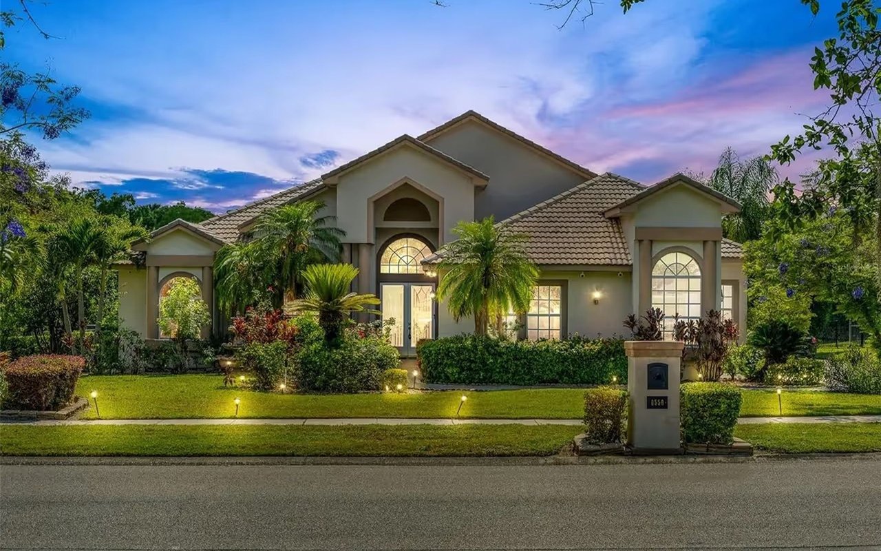 Stucco home with a tiled roof, arched windows, palms, and illuminated landscaping at dusk.