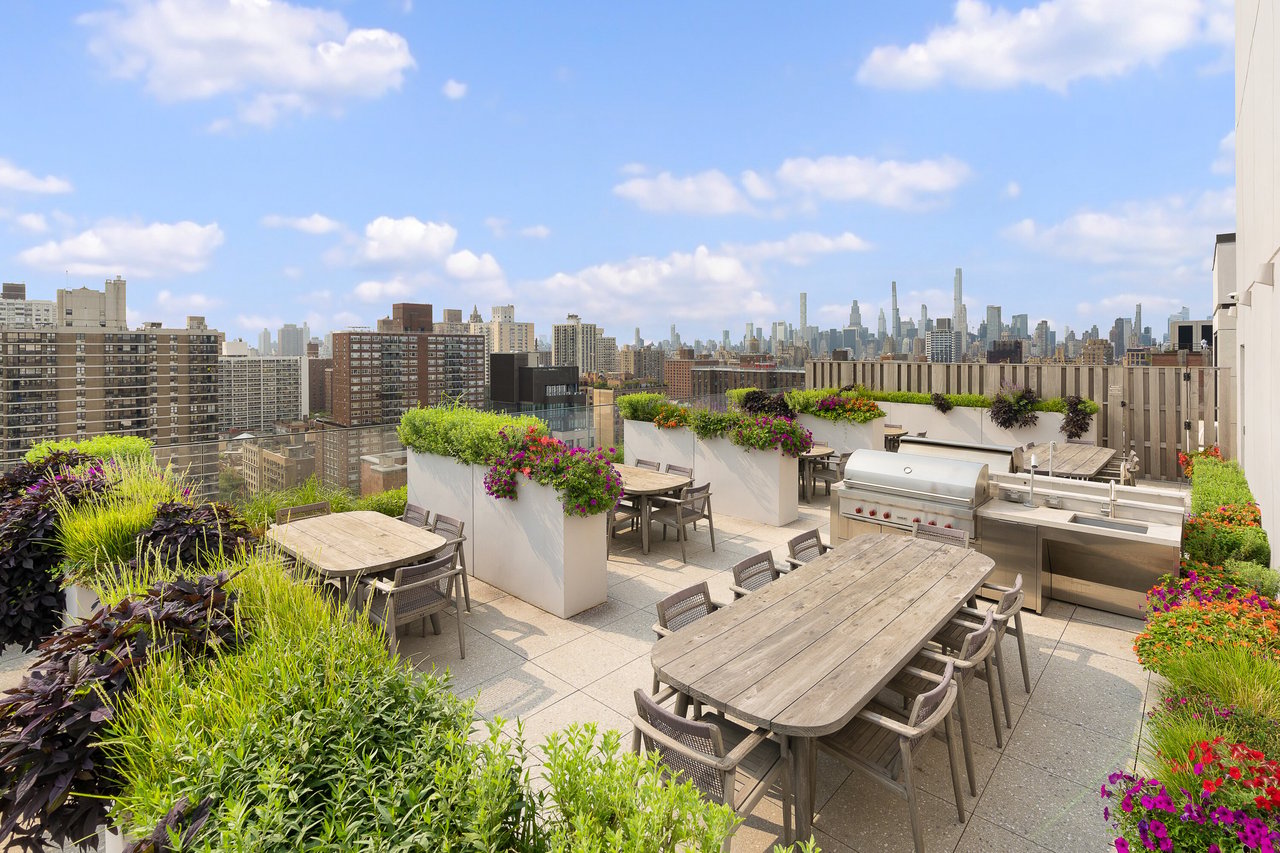Rooftop terrace with wooden dining tables, built-in grills, colorful plantings, and a Manhattan skyline view.