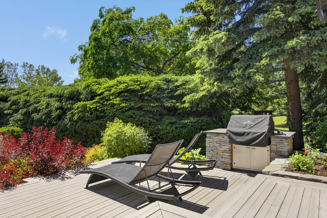 Backyard deck with woven lounge chairs and a stone outdoor kitchen beneath mature trees.