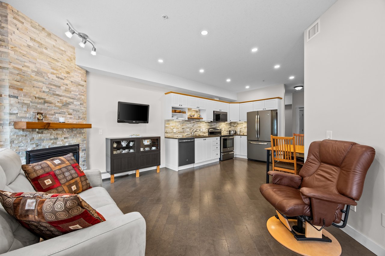Open-plan living room and kitchen with a stone fireplace wall, white cabinets, and stainless steel appliances.