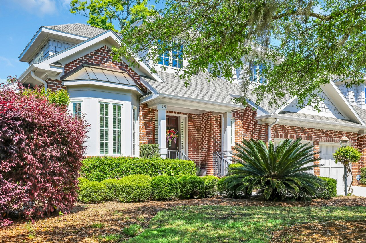 Red-brick villa with white trim, a covered entrance, clipped hedges, and a large palm in front.