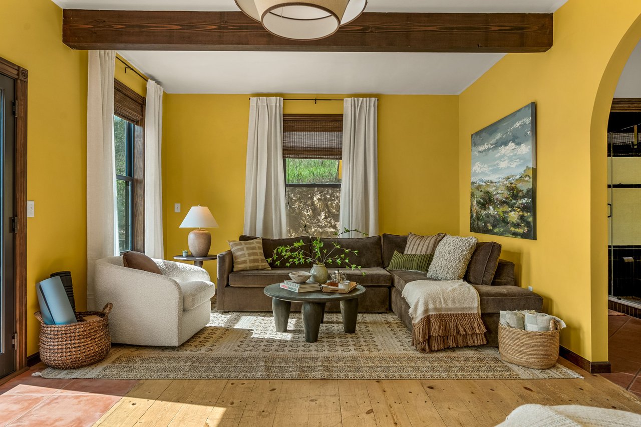 Living room with yellow walls, a brown sectional, white curtains, and an exposed wood beam.