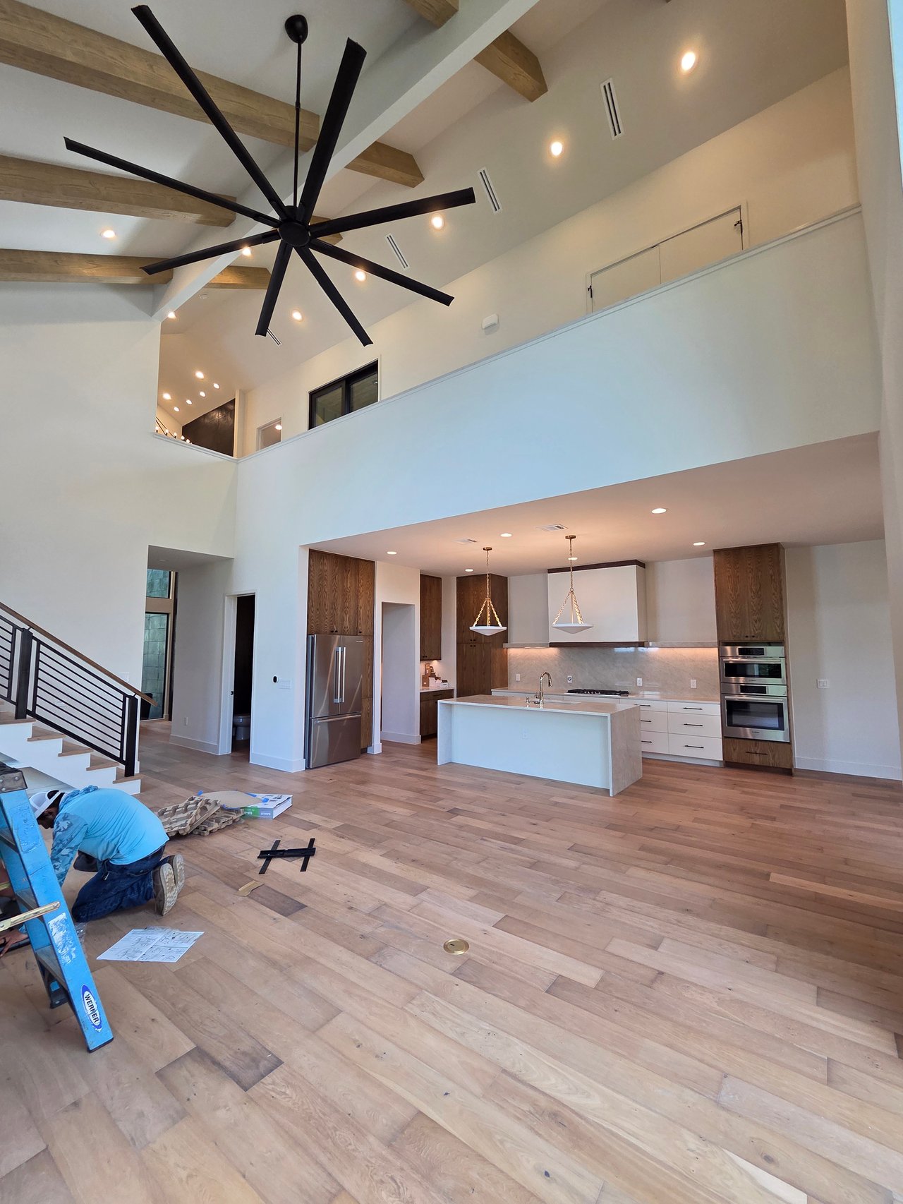 Double-height kitchen and living area with exposed wood beams, oversized ceiling fan, and a worker on the floor.