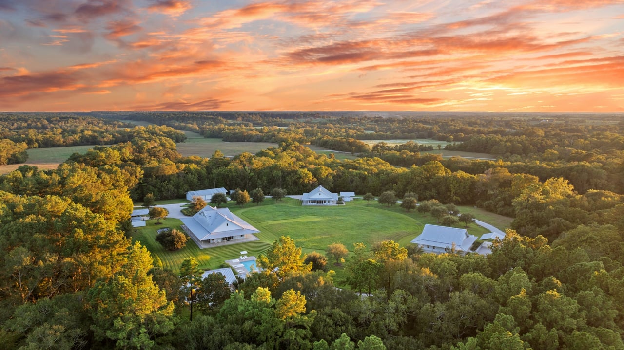 Aerial view of a rural property with several buildings, a pool, and wooded fields at sunset.