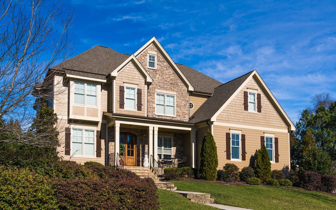 Two-story beige house with stone accents, a covered porch, and landscaped front lawn.