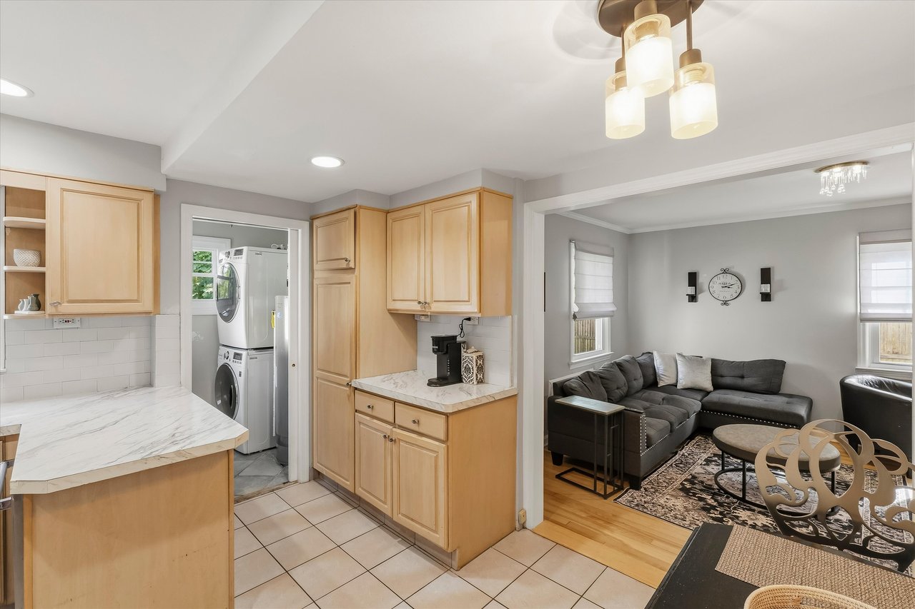 Kitchen with light wood cabinets and marble-look counters opening to a living room with a gray sectional.