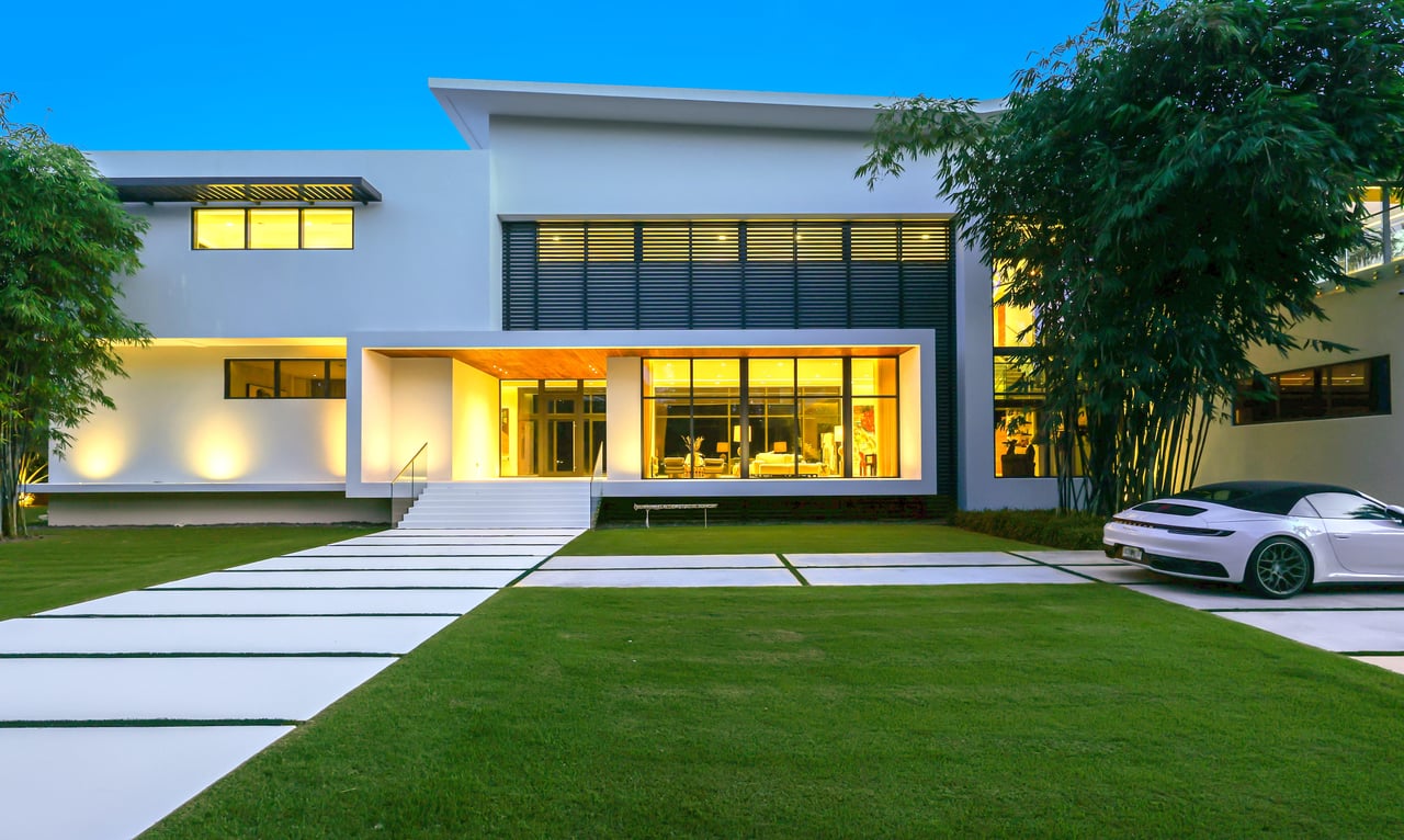 Modern white house glowing at dusk, with a geometric driveway, green lawn, and white sports car.