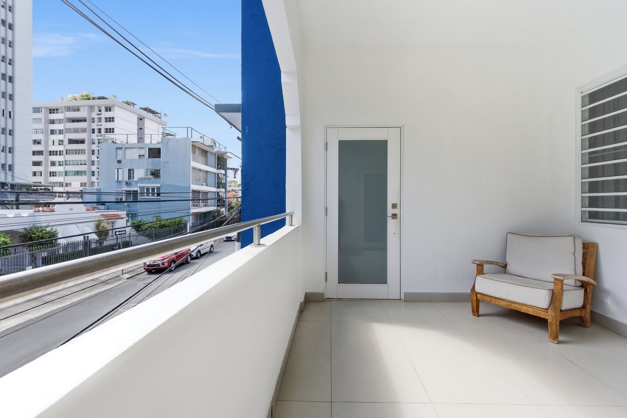 Apartment balcony with a cushioned wooden chair and a view of nearby buildings and the street.