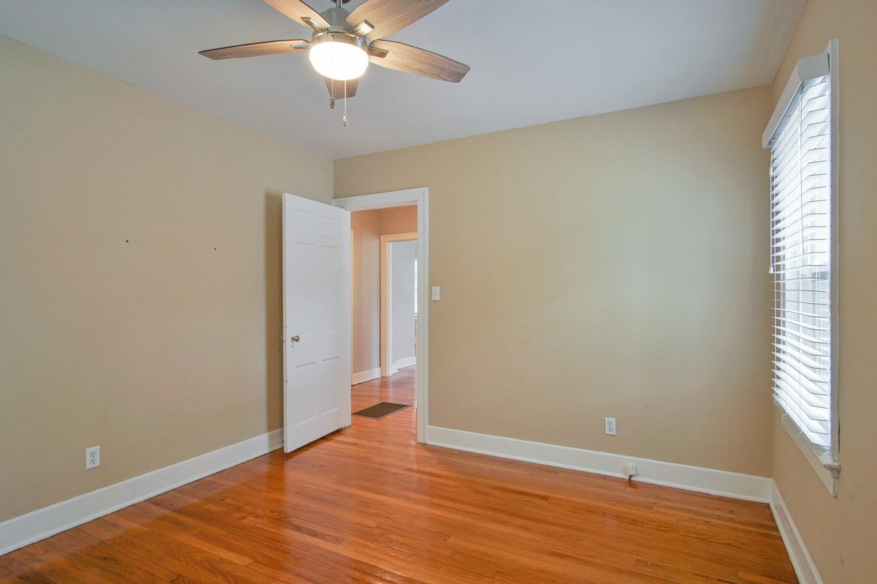 Empty room with wooden floors, beige walls, and a window with blinds. A ceiling fan with light hangs above, and an open white door leads to a hallway.