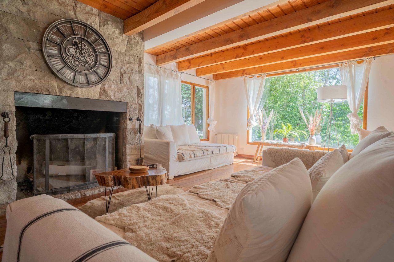 Hotel living room with a stone fireplace, exposed wood beams, white sofas, and large garden-facing windows.