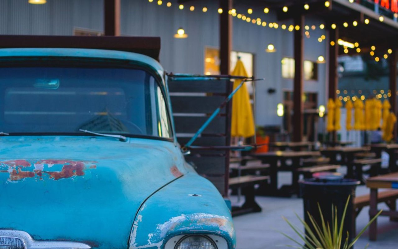 Weathered turquoise vintage pickup beside a patio dining area lit by string lights and yellow umbrellas.