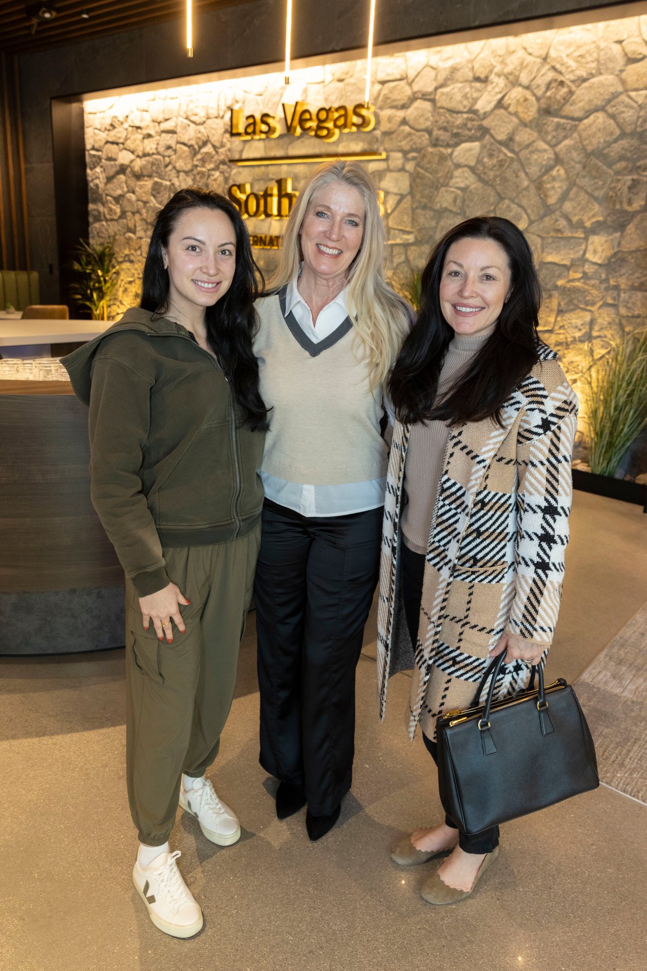Three women pose together inside a Las Vegas venue with an illuminated stone wall.