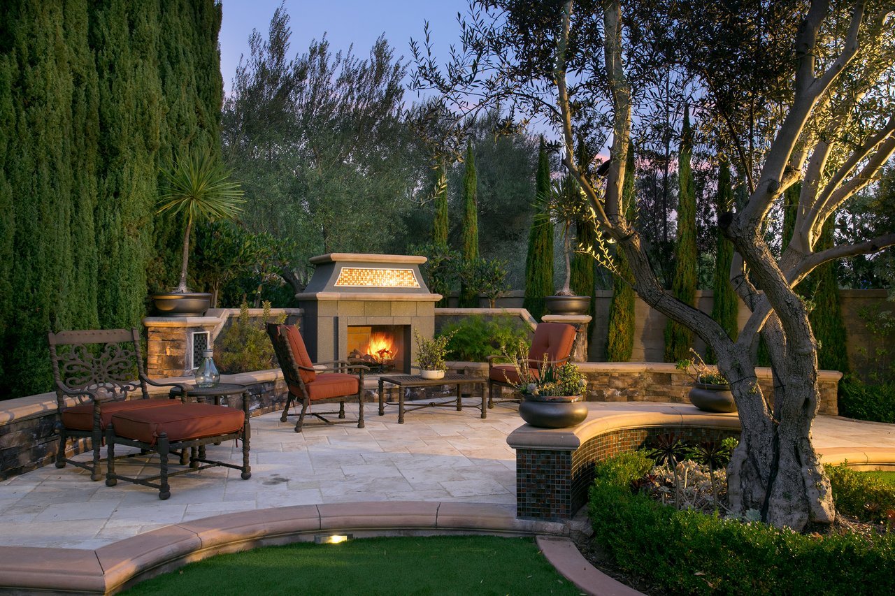 Outdoor patio with a lit stone fireplace, cushioned seating, and a curved tiled pool edge.