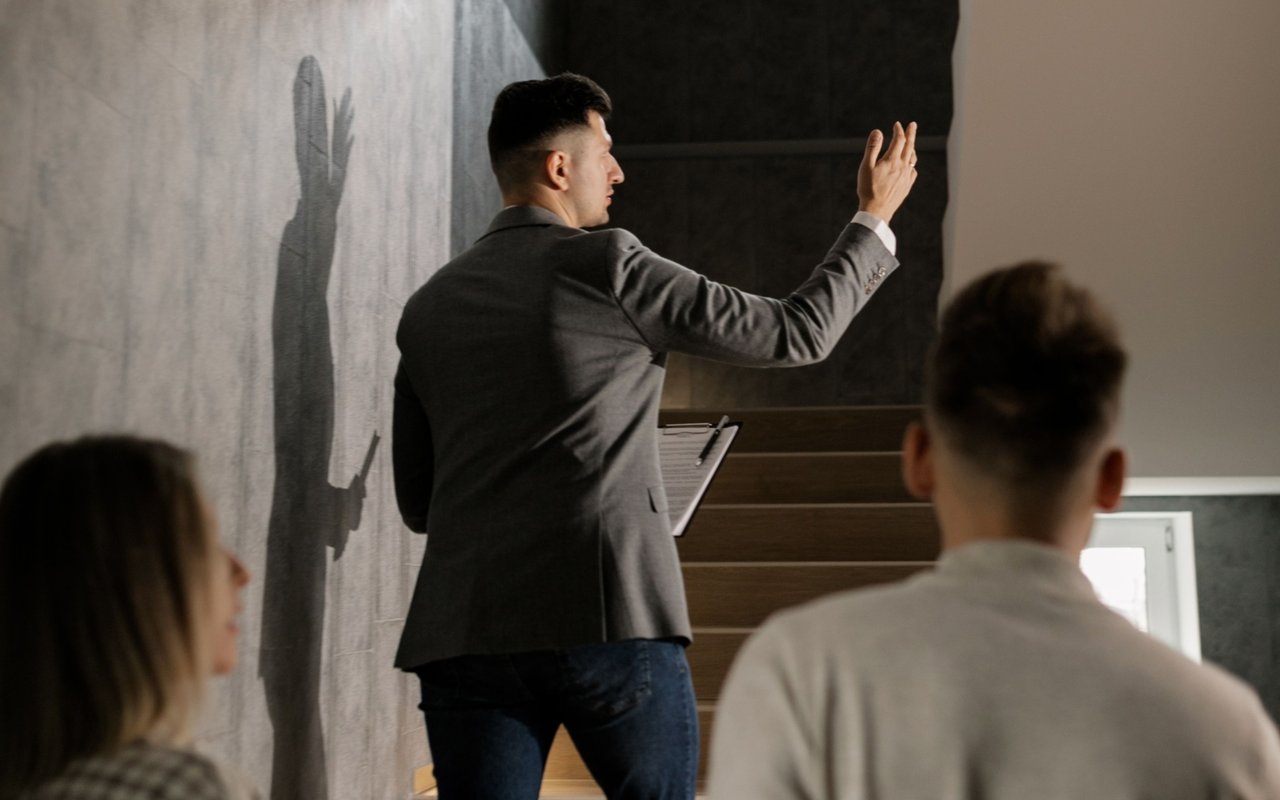 A real estate agent gestures toward a staircase while showing a home to two buyers.