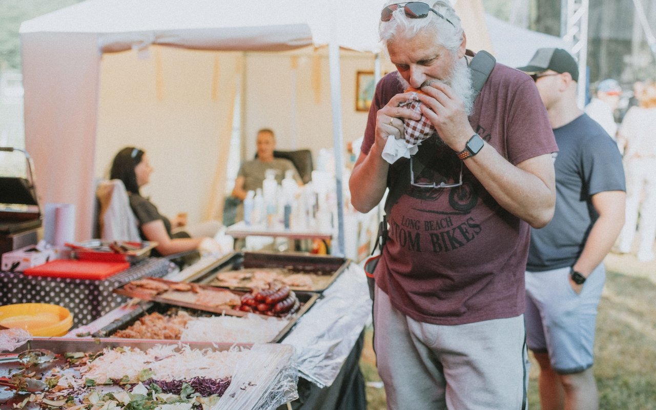 A bearded man tastes food beside a buffet of prepared dishes at an outdoor festival.