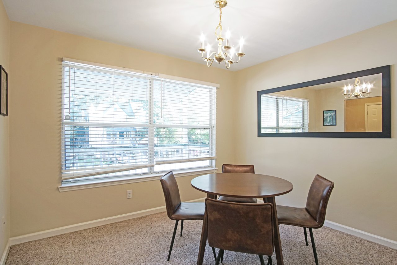 Dining room at 529 Collinsford Road, Tallahassee, Florida, featuring a round wooden table with four brown leather chairs, a large wall mirror, and a gold chandelier. The space is brightened by a large window with blinds overlooking the front porch and neighborhood.