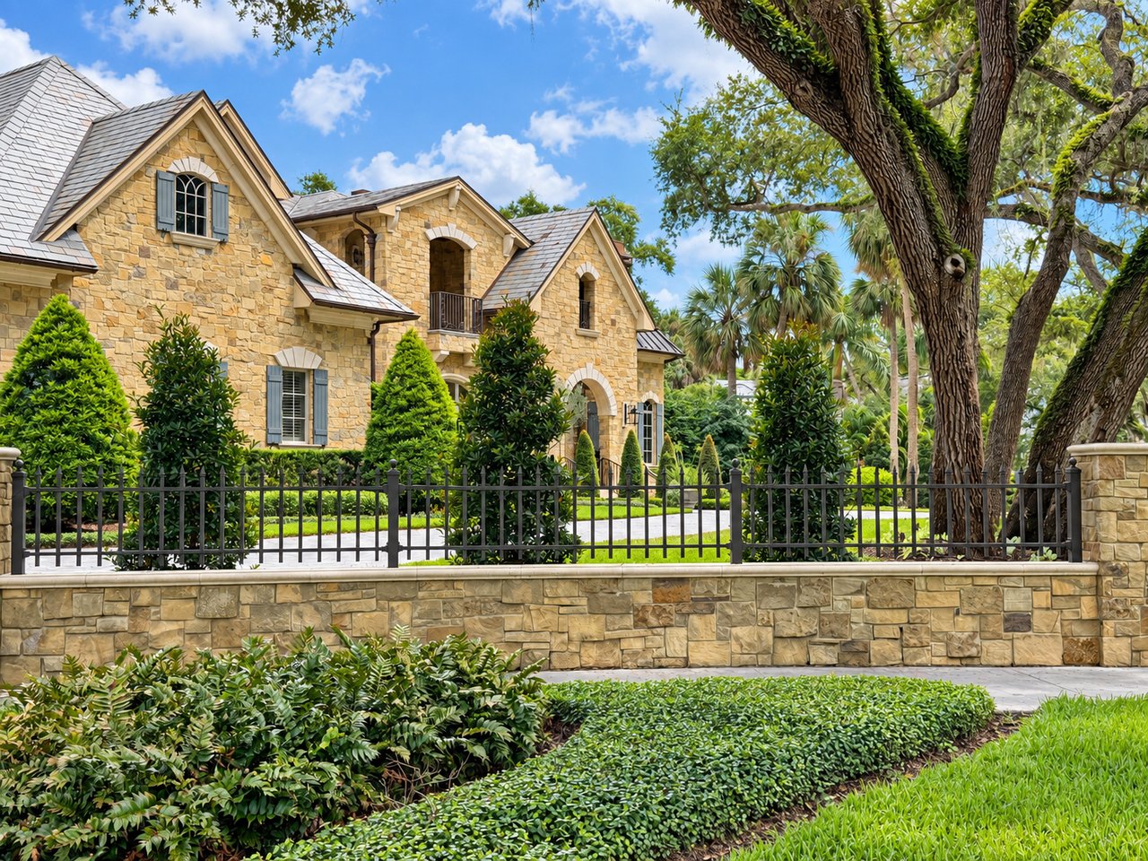 Stone house behind an iron fence and stone wall, framed by mature trees and landscaped greenery.
