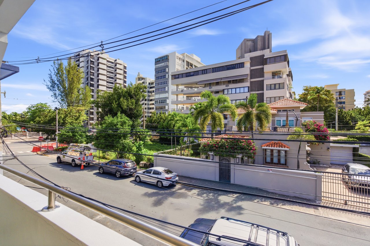 Urban apartment buildings across a street, viewed from a balcony above tropical landscaping and parked cars.