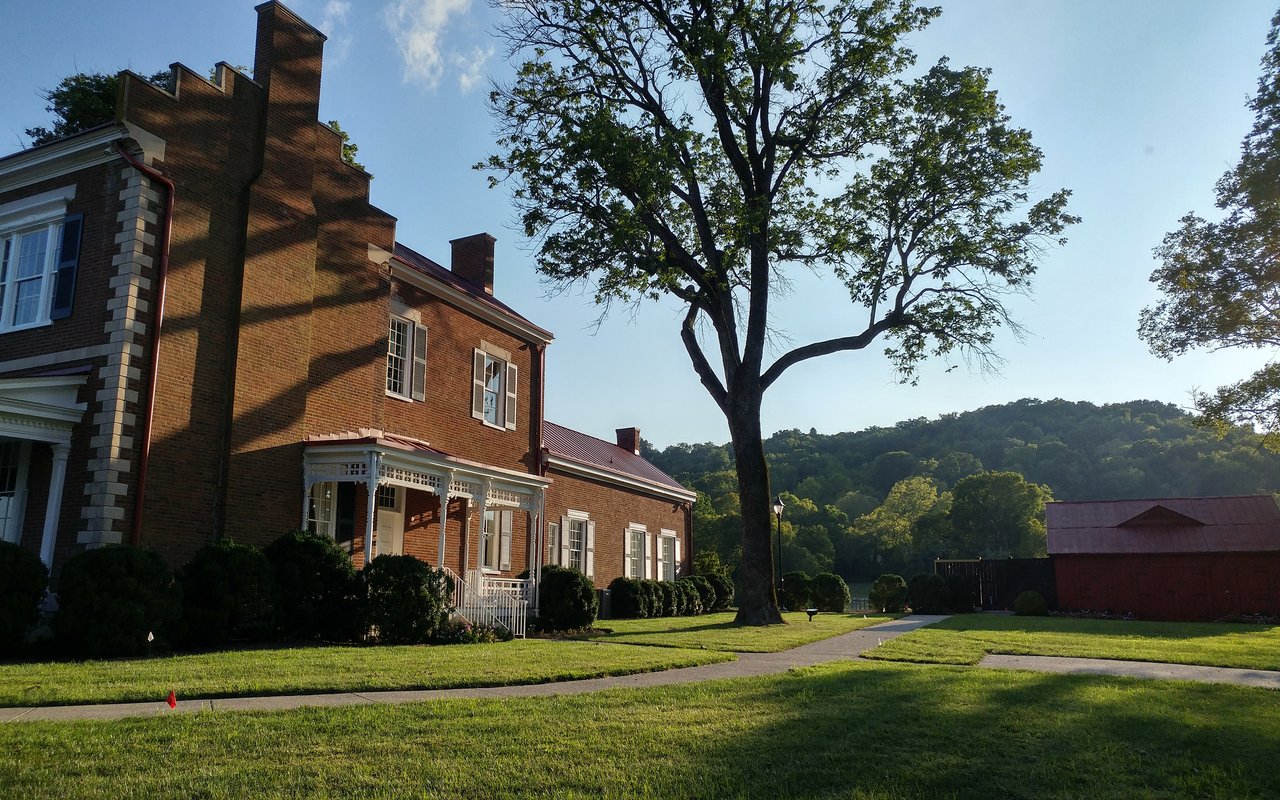 Red-brick home with a covered entrance, large shade tree, lawn, and wooded hills beyond.