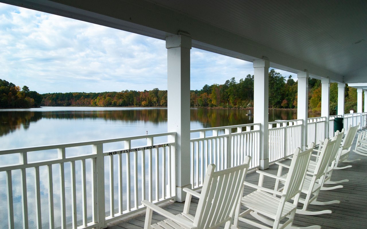 Covered lakeside deck with white rocking chairs overlooking calm water and autumn trees.