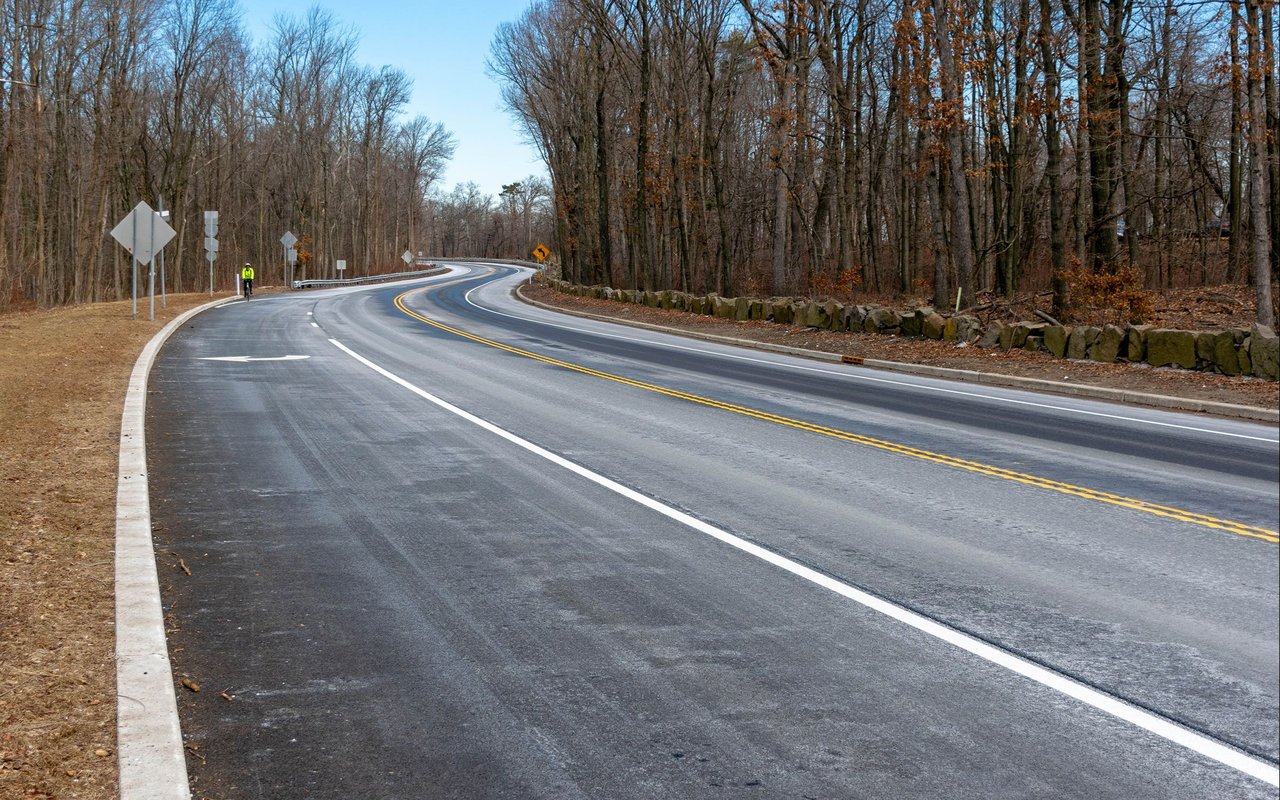 A curving road through leafless woods, with a distant person in a bright jacket.