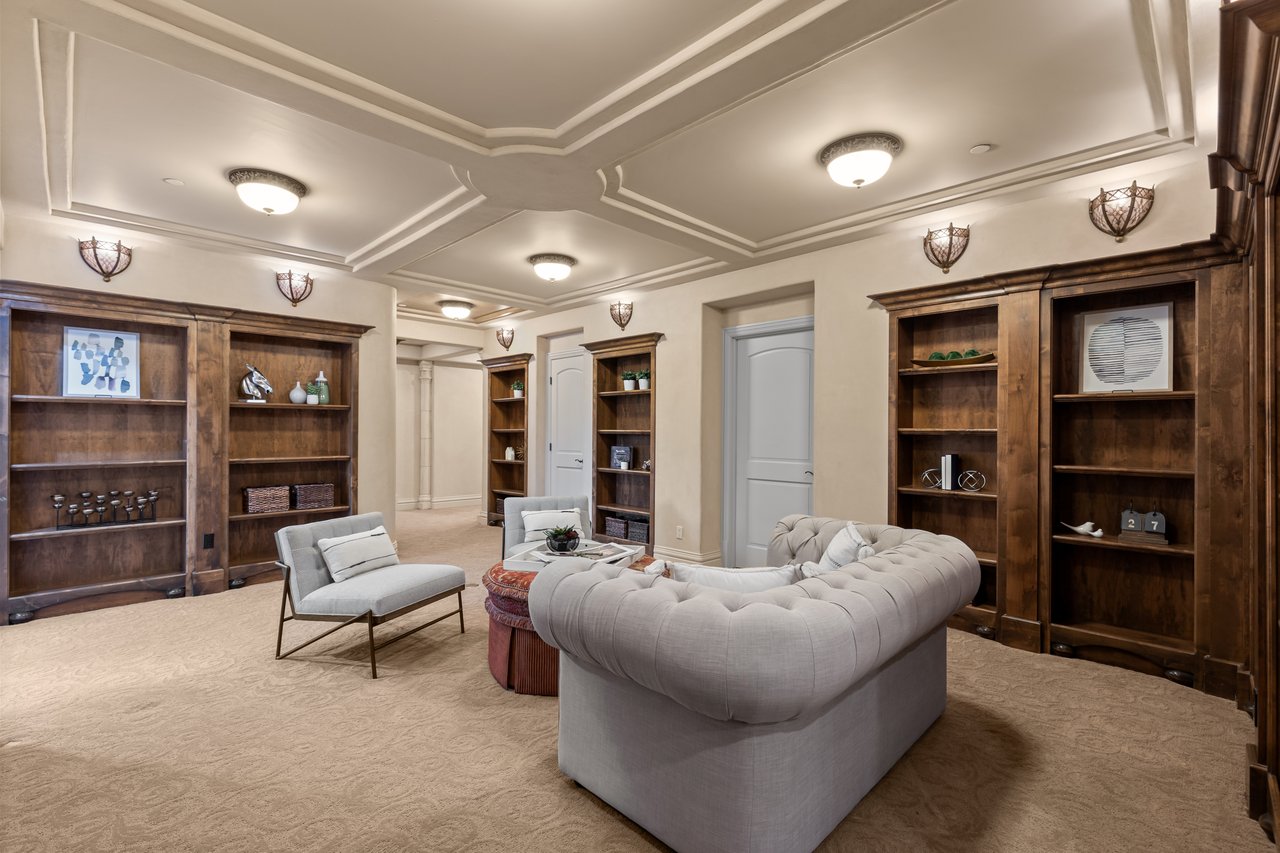 Furnished sitting room with tufted gray sofas, dark wood built-in bookshelves, and a coffered ceiling.