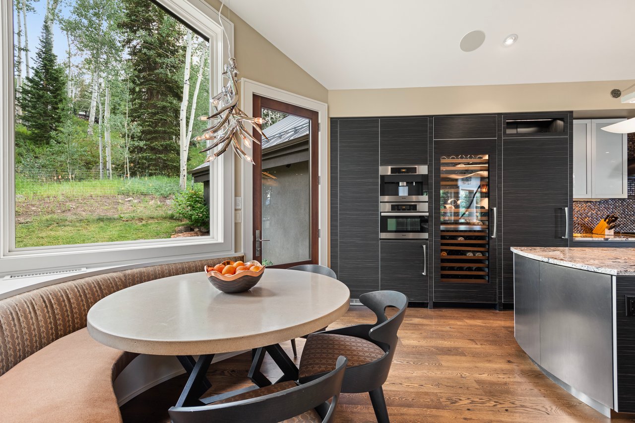 Modern kitchen dining nook with a round table, dark cabinetry, wine refrigerator, and wooded window view.