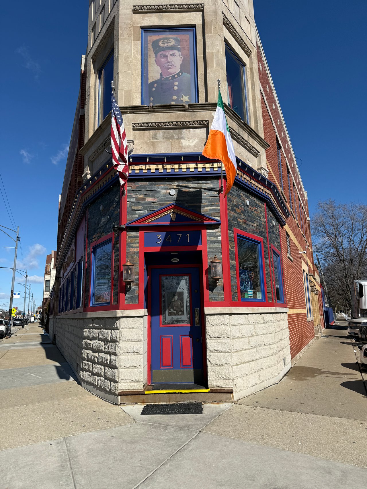 Corner storefront with red and blue trim, American and Irish flags, and a blue door numbered 3471.