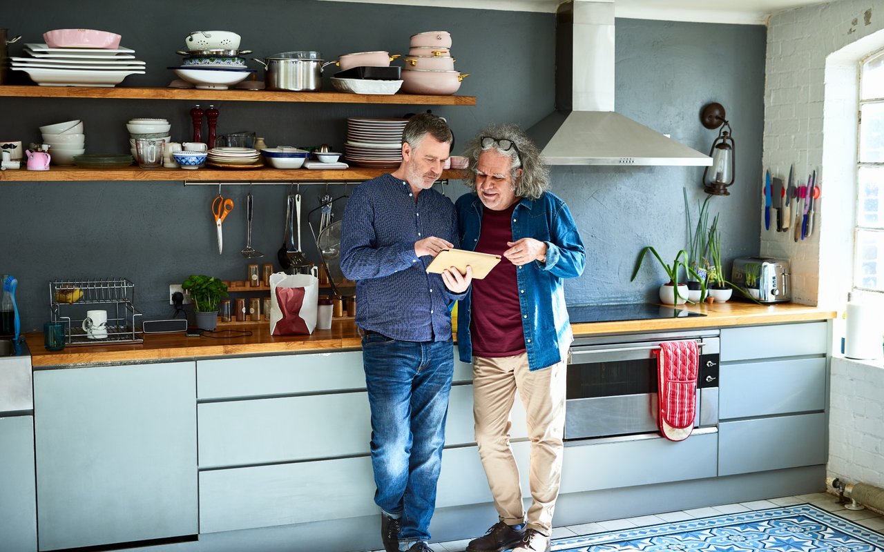 Furnished kitchen with two men reviewing a tablet beside open shelves of dishes.