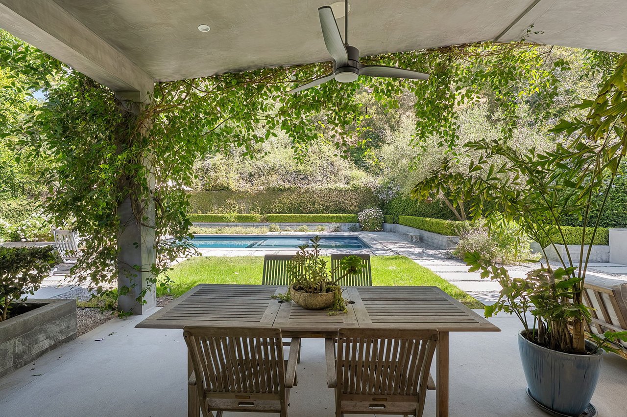Outdoor dining patio beneath a vine-covered pergola, overlooking a pool and manicured lawn.