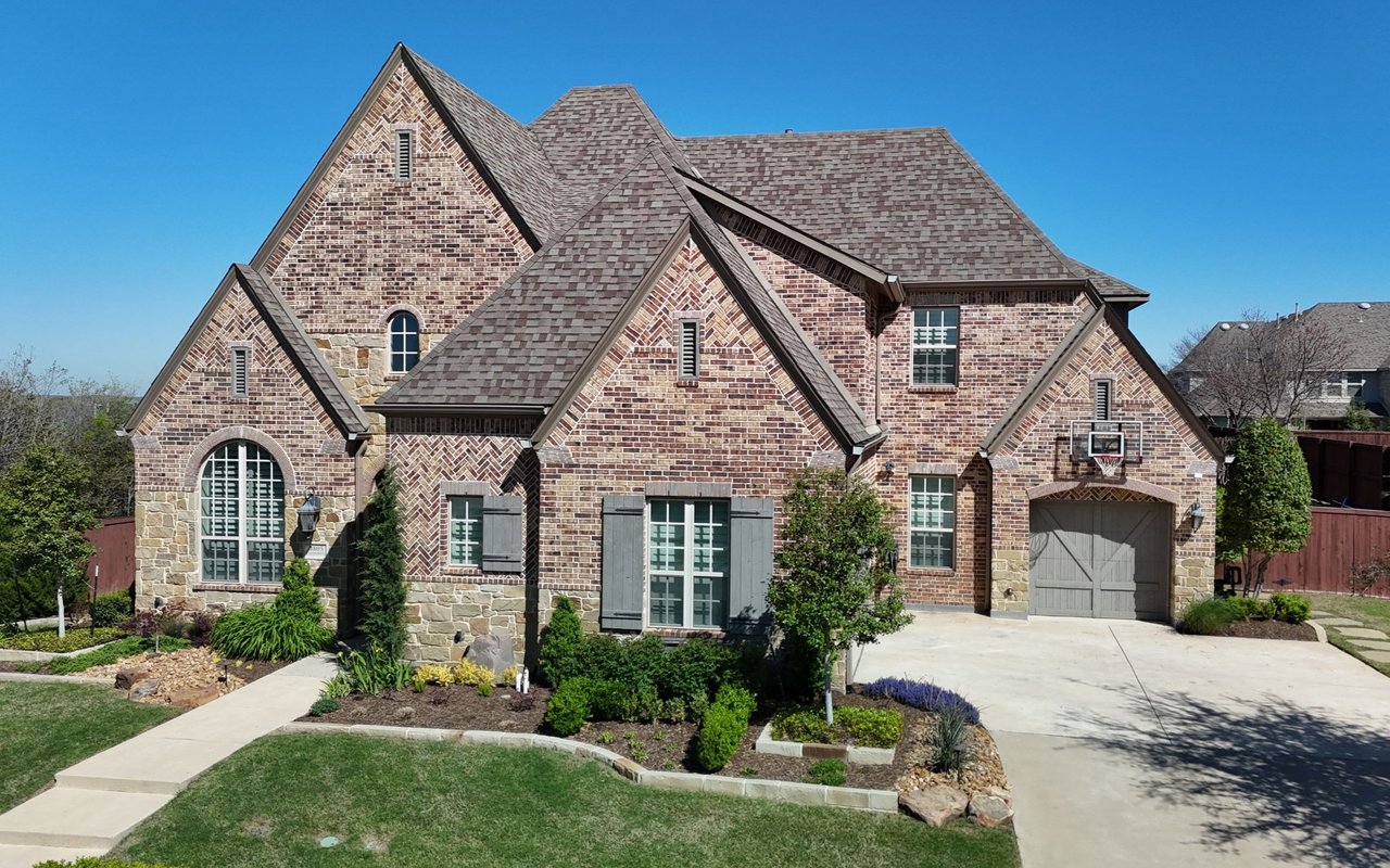 Two-story brick house with stone accents, multiple gabled roofs, landscaped yard, and attached garage.