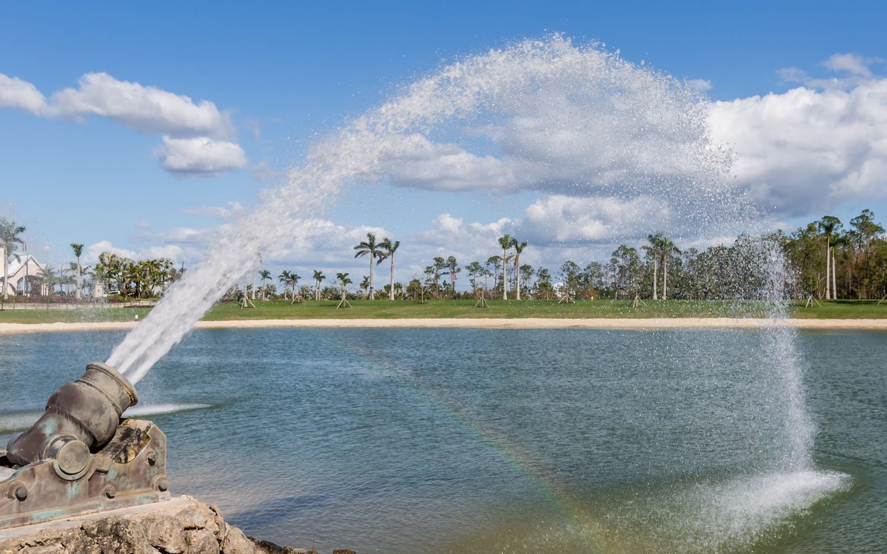 Lake fountain spraying a broad arc of water, with palm trees and homes along the far shore.