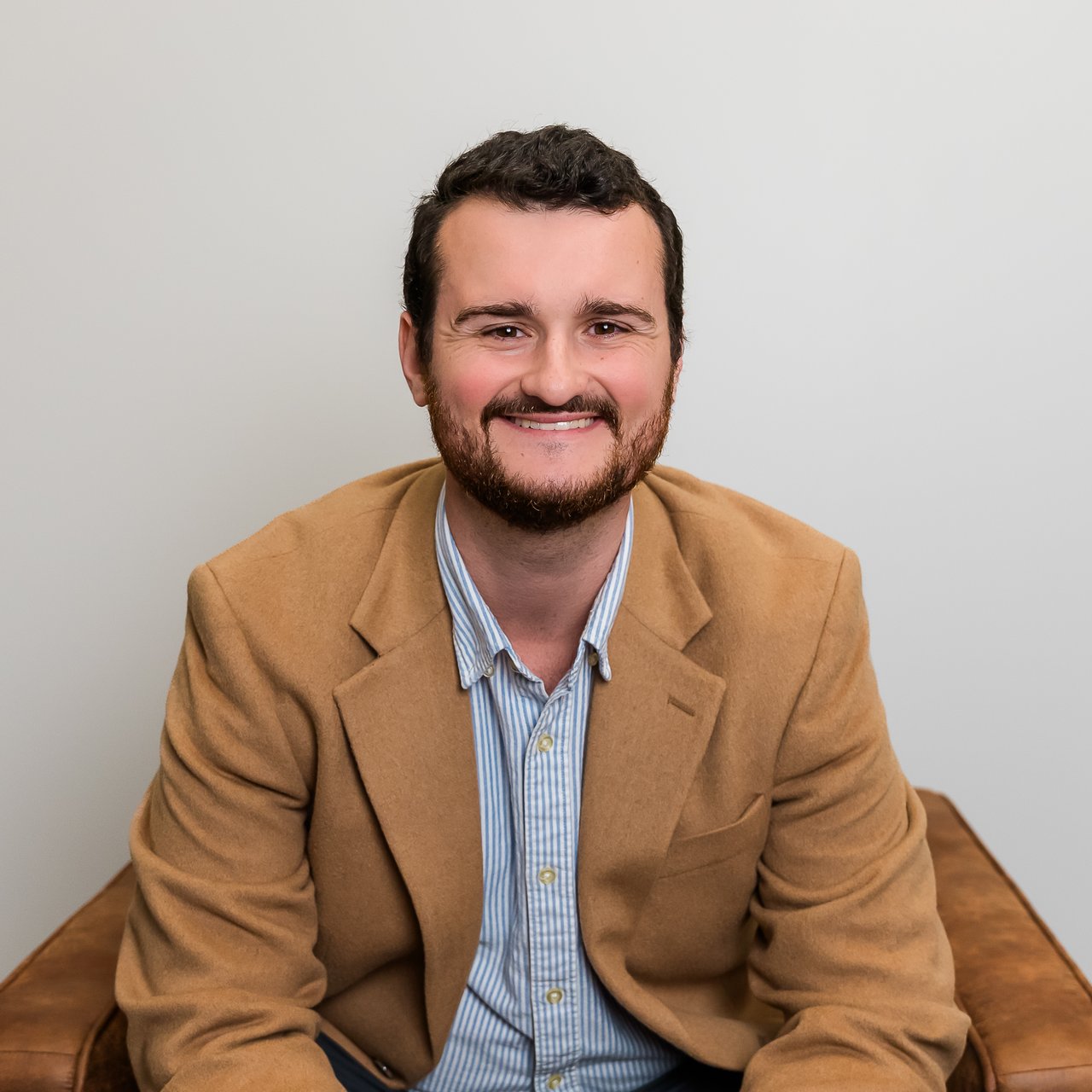 Justin Hamlin smiling in a tan blazer and blue-and-white striped shirt.