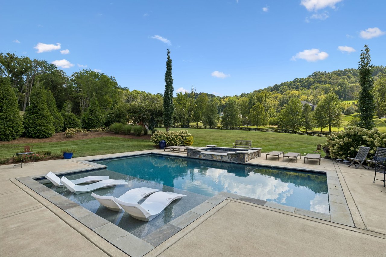 Swimming pool with white in-water loungers, stone patio, and wooded hills beyond the lawn.