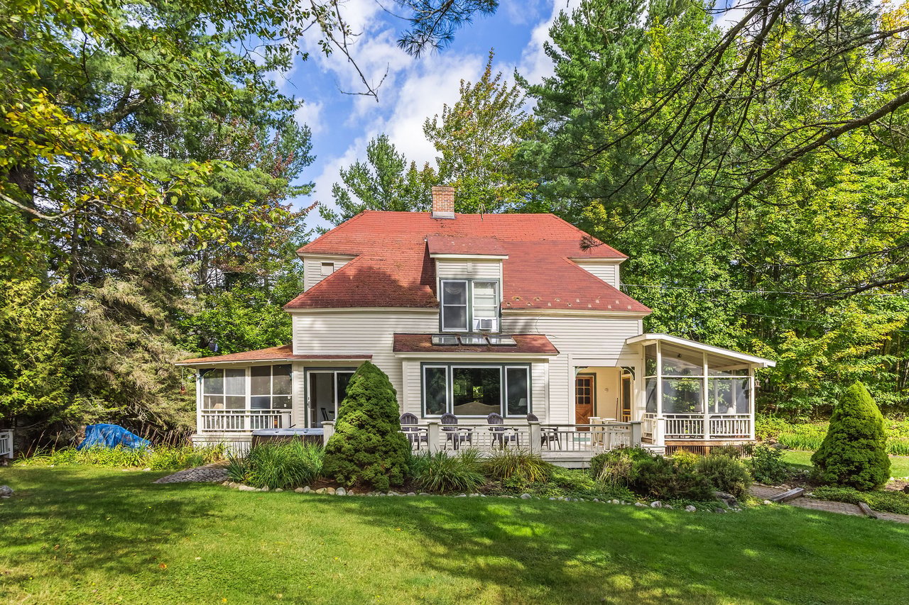 White two-story house with a red roof, screened porches, and a broad lawn beneath mature trees.
