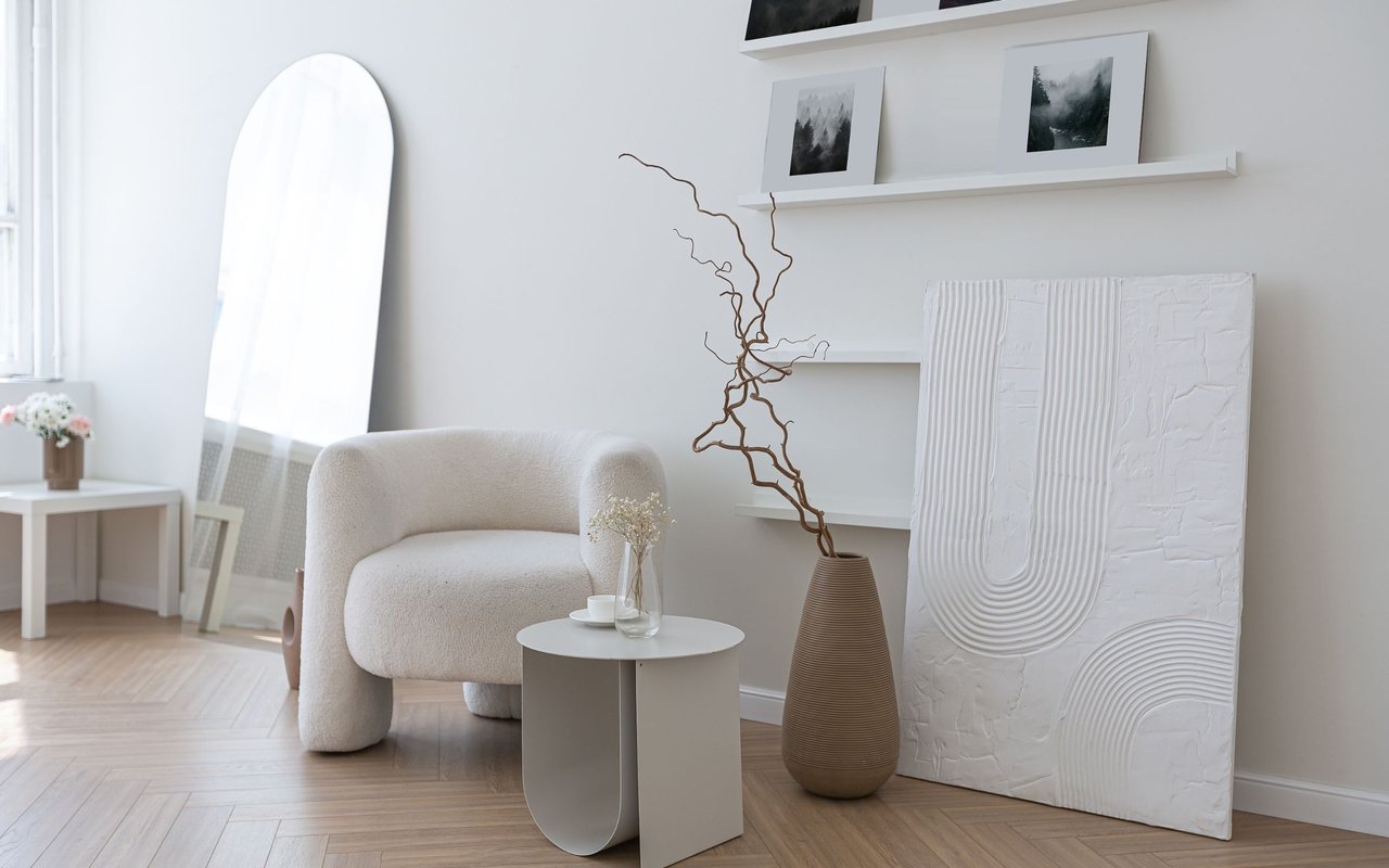 Staged living room with a cream armchair, leaning mirror, sculptural wall art, and pale herringbone flooring.