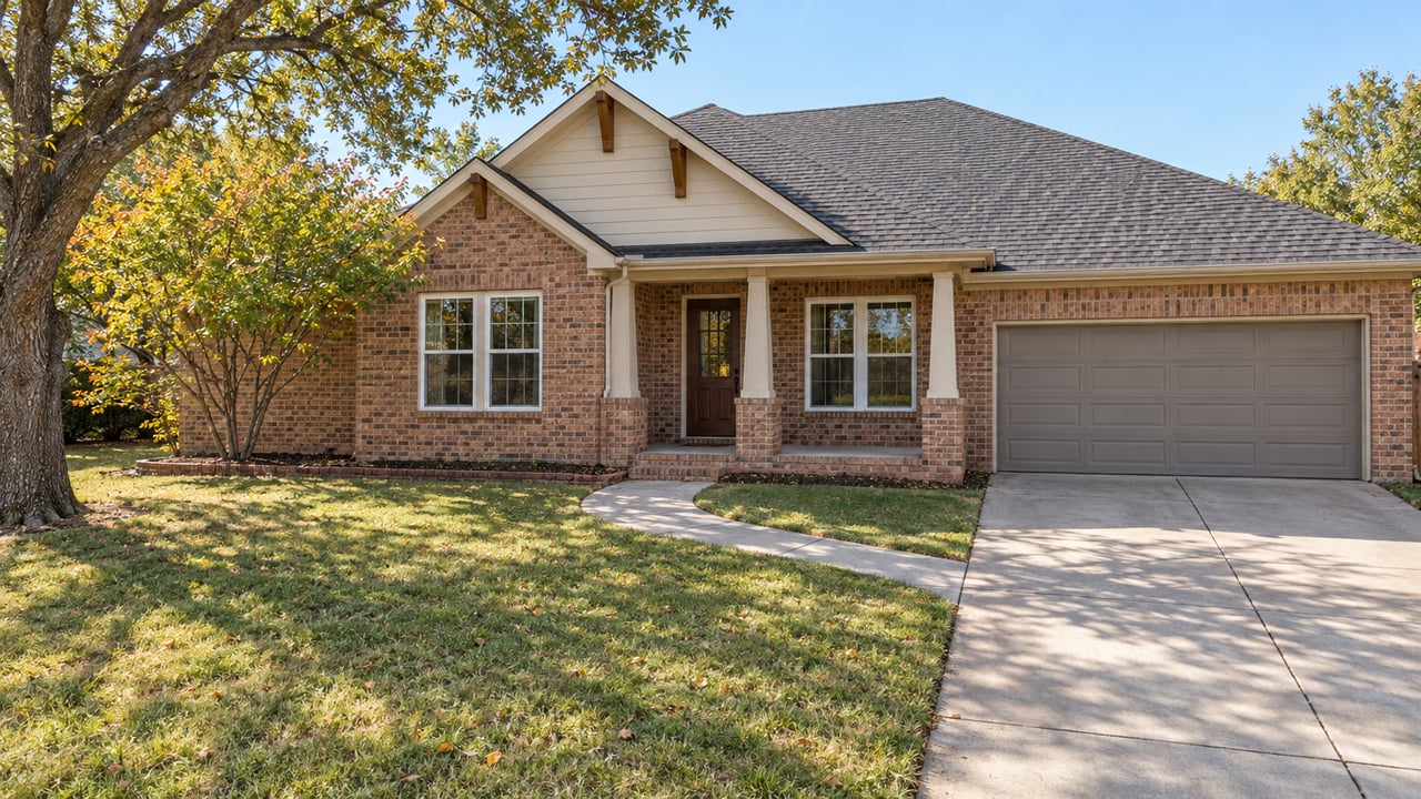 Brick single-story home with a covered porch, attached garage, deep autumn lawn and mature oak in natural daylight.