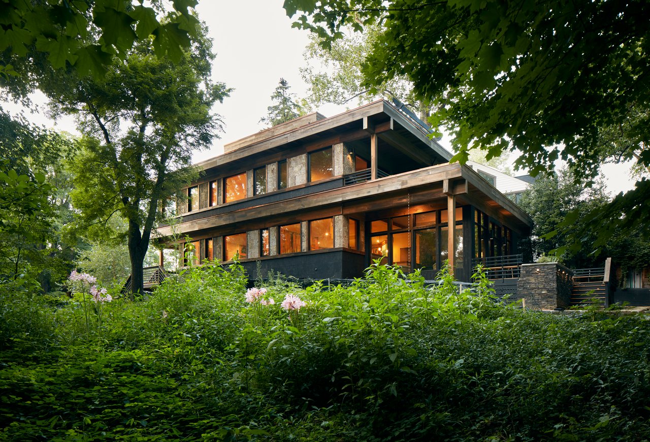 Modern two-story home with broad timber eaves and glowing windows nestled among trees and garden foliage.
