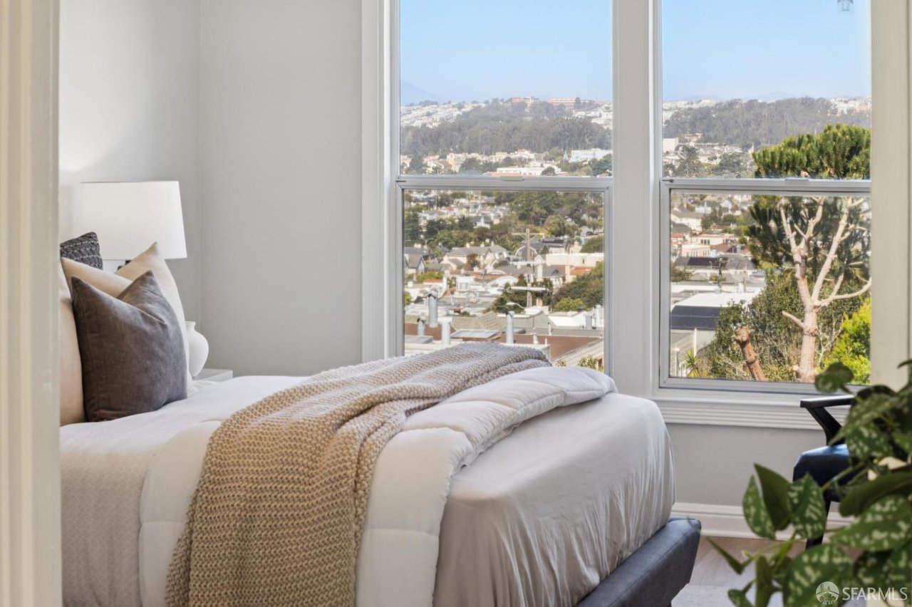 Staged bedroom with a neutral bed and tall windows overlooking a city neighborhood and hills.